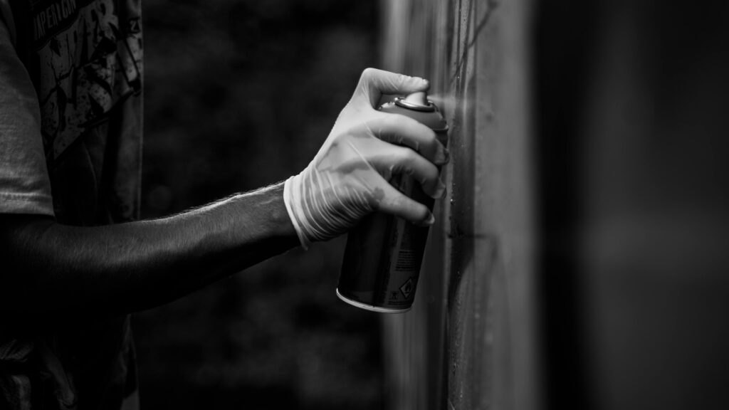 A gloved hand holding spray paint, creating graffiti art on a wall in a monochrome setting.